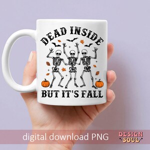 May include: White ceramic mug with a black and orange Halloween design. The mug features the text "DEAD INSIDE BUT IT'S FALL" with illustrations of skeletons, pumpkins, bats, and leaves. The mug is held by a hand.