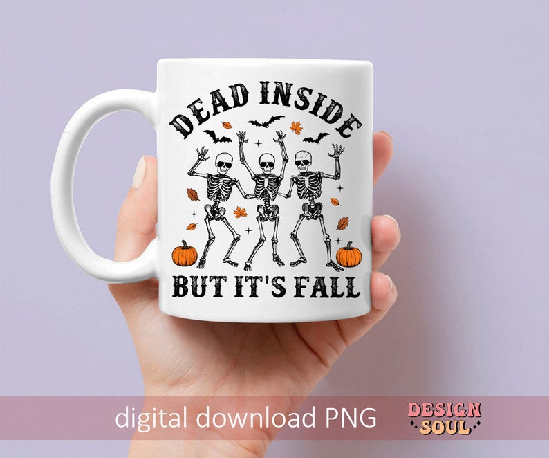 May include: White ceramic mug with a black and orange Halloween design. The mug features the text "DEAD INSIDE BUT IT'S FALL" with illustrations of skeletons, pumpkins, bats, and leaves. The mug is held by a hand.