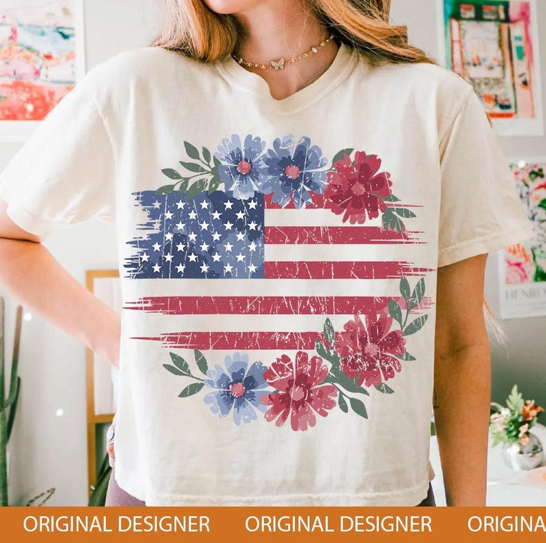 Puede incluir: Camiseta color crema con un dise&ntilde;o desgastado de la bandera estadounidense. La bandera est&aacute; adornada con una corona floral de flores azules y rojas y hojas verdes. La camiseta tiene un corte holgado.