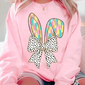 May include: A pink sweatshirt with a graphic of a white and black polka dot bow with pastel bunny ears. The bunny ears are outlined in gold and have a pastel pink, yellow, and blue pattern.