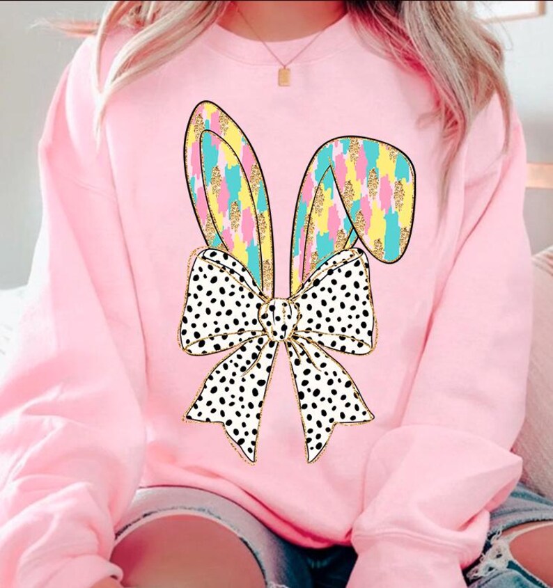 May include: A pink sweatshirt with a graphic of a white and black polka dot bow with pastel bunny ears. The bunny ears are outlined in gold and have a pastel pink, yellow, and blue pattern.
