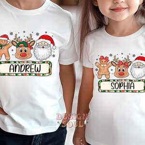 May include: Two white t-shirts with Christmas themed graphics. The first shirt has a gingerbread man, reindeer, and Santa Claus with the name "Andrew" printed below. The second shirt has a gingerbread woman, reindeer, and Santa Claus with the name "Sophia" printed below.