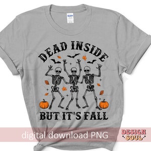 May include: A light gray t-shirt featuring a Halloween design. The graphic includes three dancing skeletons, pumpkins, bats, and fall leaves. The text reads "DEAD INSIDE BUT IT'S FALL".