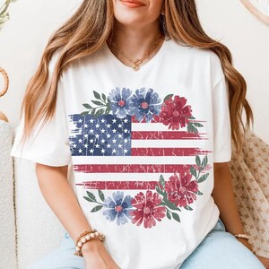 Puede incluir: Camiseta blanca con un dise&ntilde;o desgastado de la bandera estadounidense, con elementos florales rojos y azules. El dise&ntilde;o de la bandera incluye estrellas y rayas, rodeado por una corona floral. Un estilo casual y patri&oacute;tico.