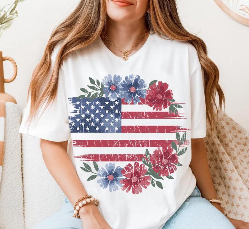 Puede incluir: Camiseta blanca con un dise&ntilde;o desgastado de la bandera estadounidense, con elementos florales rojos y azules. El dise&ntilde;o de la bandera incluye estrellas y rayas, rodeado por una corona floral. Un estilo casual y patri&oacute;tico.