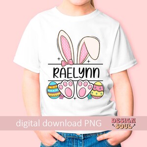 May include: White t-shirt with a pink bunny ears design, a pink bow, and the name "Raelynn" printed on the front. The shirt also features colorful Easter eggs and paw prints.