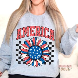 May include: A light gray sweatshirt with the word "AMERICA" printed in red and blue letters. A red, white, and blue sunflower with a checkered background is printed below the word.
