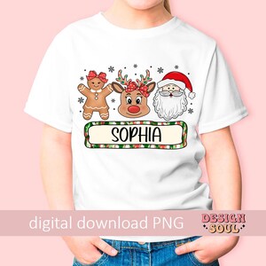 May include: White t-shirt with a Christmas design featuring a gingerbread person, a reindeer, and Santa Claus. The design includes the name "Sophia" in a green and red plaid banner.