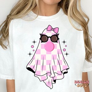 May include: White t-shirt with a pink and white checkered ghost wearing black sunglasses and a pink bow. The ghost has a pink bubble gum in its mouth. The text "DESIGN SOUL" is in the bottom right corner of the shirt.