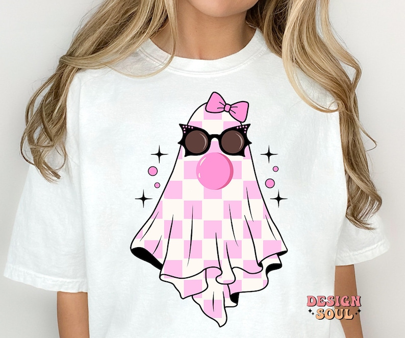 May include: White t-shirt with a pink and white checkered ghost wearing black sunglasses and a pink bow. The ghost has a pink bubble gum in its mouth. The text "DESIGN SOUL" is in the bottom right corner of the shirt.