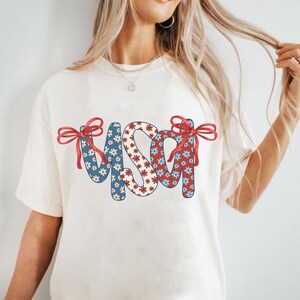 May include: White t-shirt with a red, white, and blue floral design that spells out "USA" with red bows on each side.
