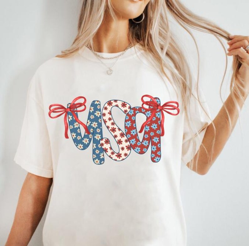 May include: White t-shirt with a red, white, and blue floral design that spells out "USA" with red bows on each side.