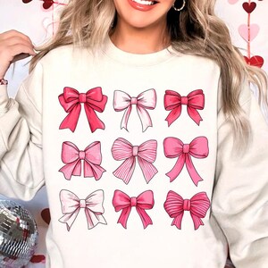 May include: A white sweatshirt with a pink and white bow pattern. The bows are arranged in a grid of nine.