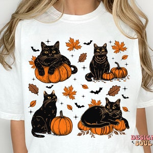 May include: White t-shirt featuring a Halloween design with four black cats, pumpkins, bats, and autumn leaves. The cats are in various poses, some sitting on pumpkins. The design has orange, black, and brown colors.