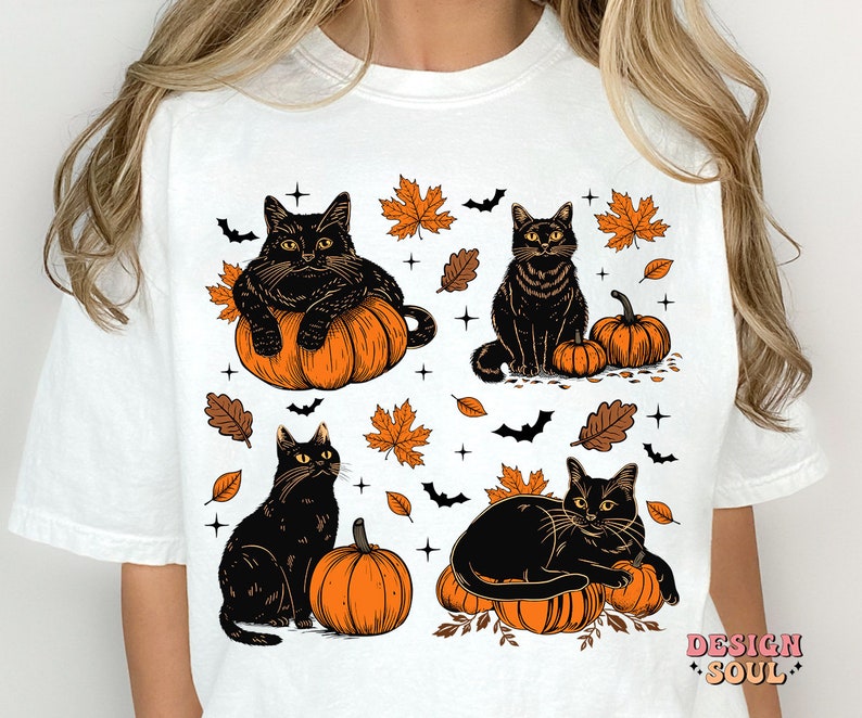May include: White t-shirt featuring a Halloween design with four black cats, pumpkins, bats, and autumn leaves. The cats are in various poses, some sitting on pumpkins. The design has orange, black, and brown colors.