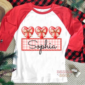 May include: A white and red baseball tee shirt with three red hearts with bows and a gingham banner with the name "Sophia" printed on it.