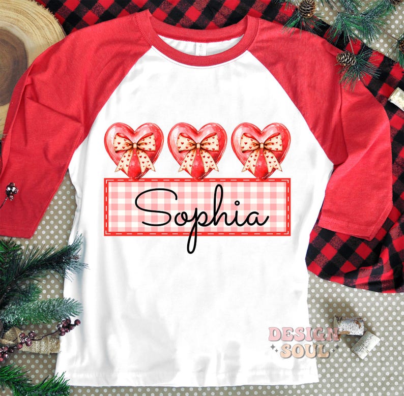 May include: A white and red baseball tee shirt with three red hearts with bows and a gingham banner with the name "Sophia" printed on it.