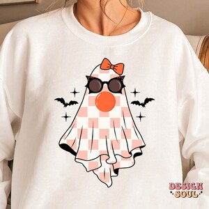 May include: White sweatshirt with a pink and white checkered ghost wearing sunglasses and a red bow. The ghost has a red nose and is surrounded by stars and bats.