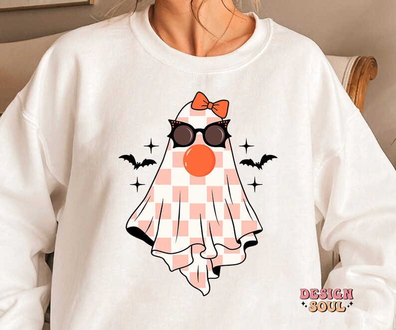 May include: White sweatshirt with a pink and white checkered ghost wearing sunglasses and a red bow. The ghost has a red nose and is surrounded by stars and bats.