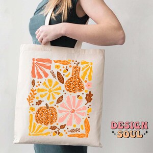 May include: A white tote bag with a colorful design featuring pumpkins, flowers, and leaves. The design is in a retro style with a mix of orange, yellow, pink, and brown colors. The text "DESIGN SOUL" is printed in pink on the bottom right corner of the bag.