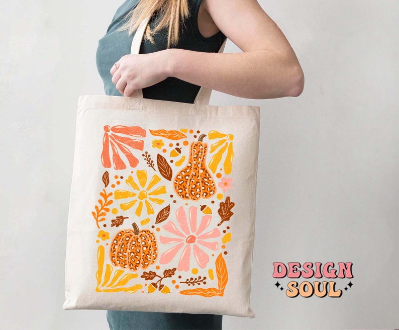 May include: A white tote bag with a colorful design featuring pumpkins, flowers, and leaves. The design is in a retro style with a mix of orange, yellow, pink, and brown colors. The text "DESIGN SOUL" is printed in pink on the bottom right corner of the bag.