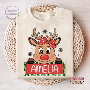 May include: A beige Christmas-themed sweatshirt with a cartoon reindeer wearing a red bow. The reindeer is peeking over a green and white striped banner with the name "Amelia" printed in red.