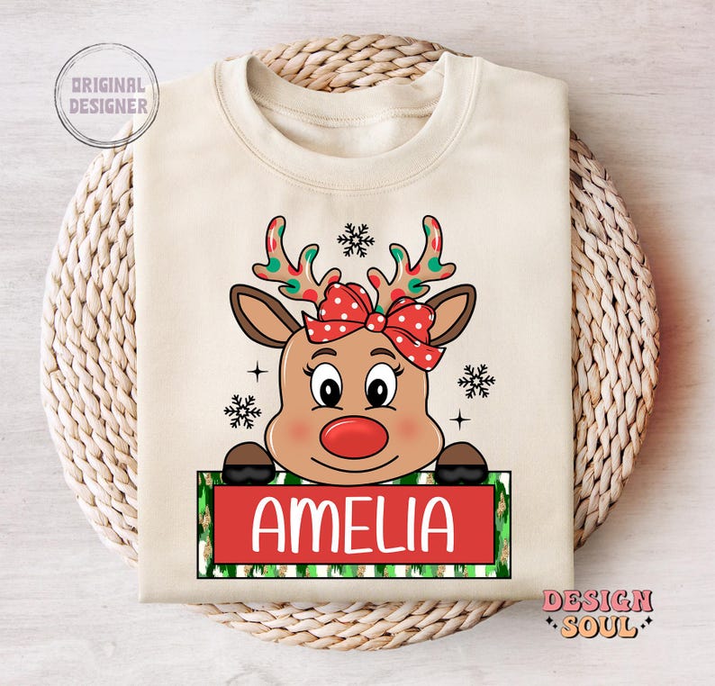 May include: A beige Christmas-themed sweatshirt with a cartoon reindeer wearing a red bow. The reindeer is peeking over a green and white striped banner with the name "Amelia" printed in red.