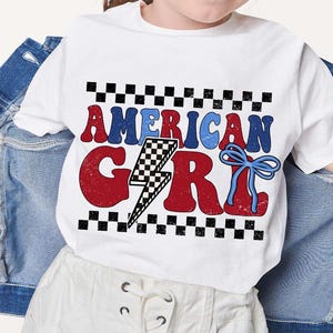 May include: White t-shirt with a red, white, and blue graphic that says "American Girl" with a lightning bolt and a blue bow. The graphic is surrounded by a black and white checkered pattern.