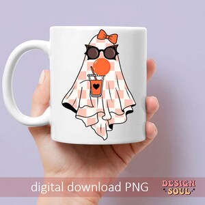 May include: White ceramic mug with a pink and white checkered ghost wearing sunglasses and holding a glass of orange juice. The ghost has a pink bow on its head. The mug has the text "Design Soul" on the bottom.
