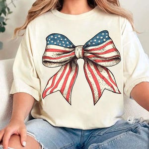 Peut inclure: Un t-shirt blanc avec un n&oelig;ud rouge, blanc et bleu fait d'un imprim&eacute; de drapeau am&eacute;ricain.
