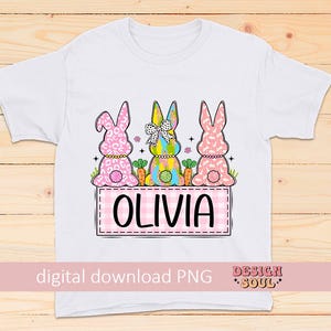 May include: White t-shirt with a pink, yellow, and blue bunny design. The bunnies are wearing floral and polka dot patterns. The text "Olivia" is printed on a pink and white checkered background.