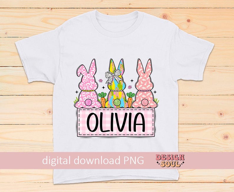 May include: White t-shirt with a pink, yellow, and blue bunny design. The bunnies are wearing floral and polka dot patterns. The text "Olivia" is printed on a pink and white checkered background.