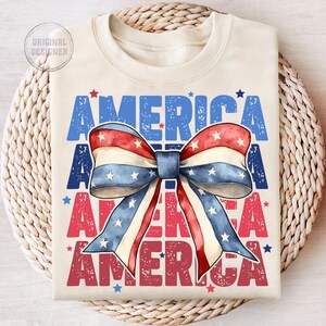 May include: Cream-colored t-shirt with the word "AMERICA" in blue and red, and a watercolor bow in red, white, and blue. The bow has white stars. The shirt is folded on a woven surface.