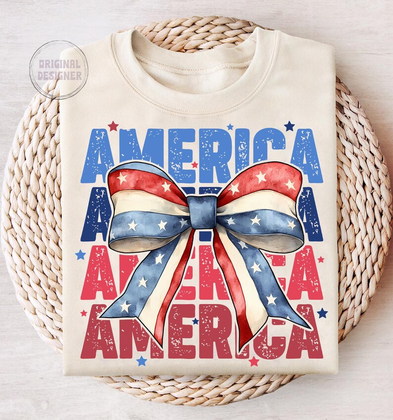 May include: Cream-colored t-shirt with the word "AMERICA" in blue and red, and a watercolor bow in red, white, and blue. The bow has white stars. The shirt is folded on a woven surface.