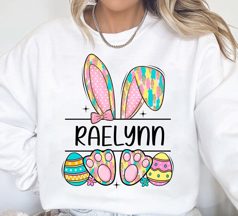 May include: White sweatshirt with a pink and yellow bunny ear design, the name "RAELynn" in black text, and colorful Easter eggs.