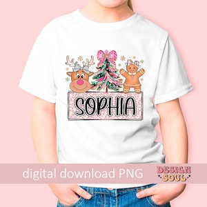 Op de afbeelding: Een wit T-shirt met een roze, groene en bruine kerstontwerp met een peperkoekman, een rendier en een kerstboom. Het ontwerp bevat ook de naam "Sophia" in roze letters.