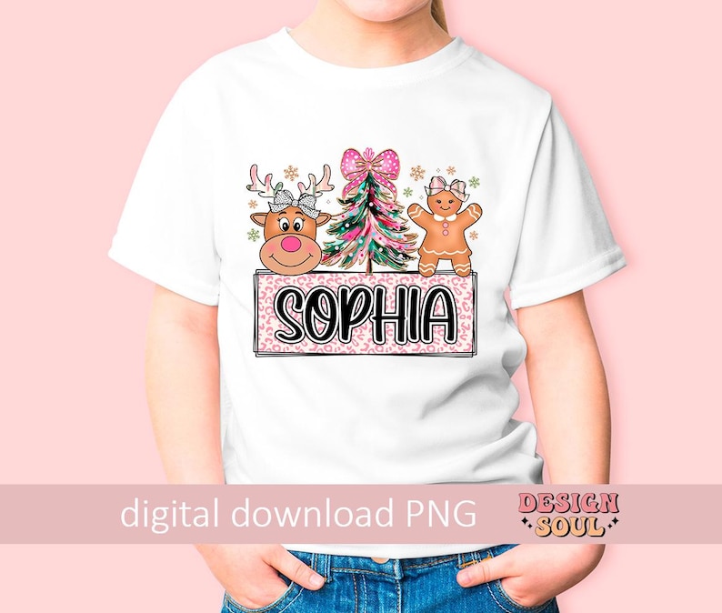 Op de afbeelding: Een wit T-shirt met een roze, groene en bruine kerstontwerp met een peperkoekman, een rendier en een kerstboom. Het ontwerp bevat ook de naam "Sophia" in roze letters.