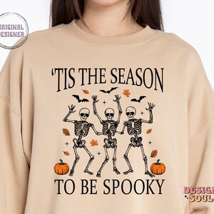 May include: Tan sweatshirt with a graphic of three skeletons dancing with the text 'Tis the season to be spooky' and two pumpkins on either side.