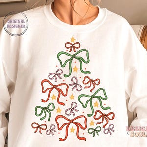 May include: White sweatshirt featuring a Christmas tree design made from green, red and pink bows. The bows are decorated with gold stars and red dots.
