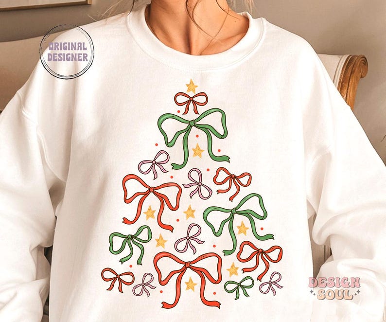 May include: White sweatshirt featuring a Christmas tree design made from green, red and pink bows. The bows are decorated with gold stars and red dots.