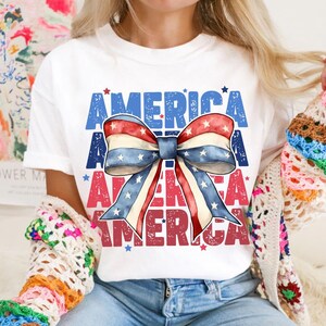 May include: White t-shirt featuring a patriotic bow design with red, white, and blue stripes and stars. The word "AMERICA" is printed in blue and red, creating a festive and eye-catching graphic.