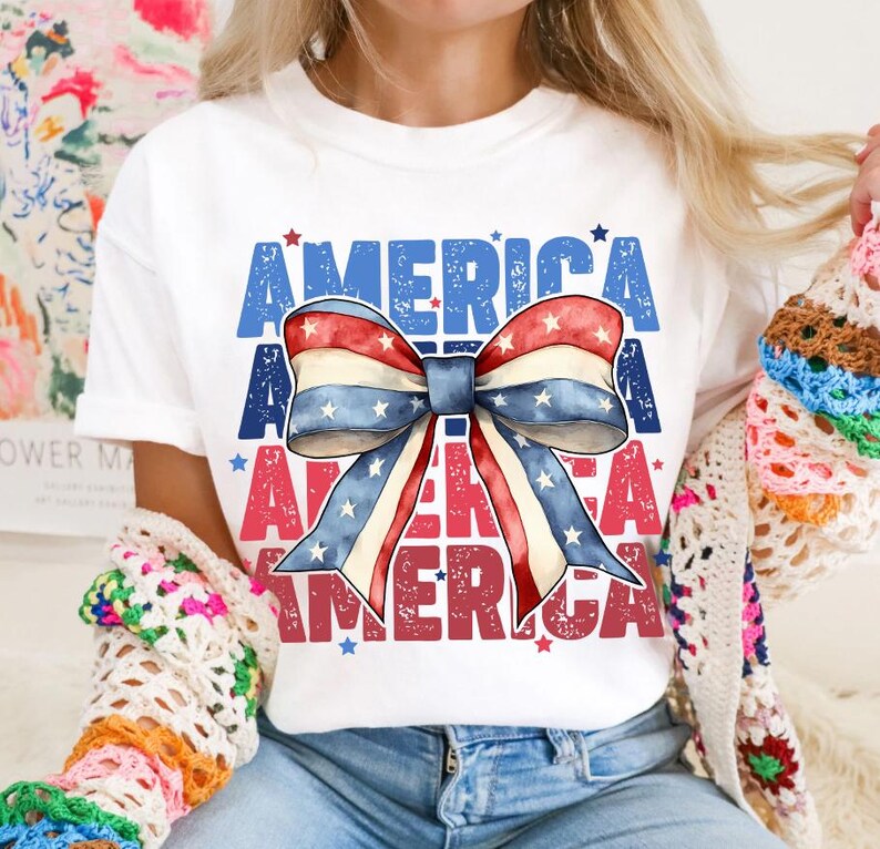 May include: White t-shirt featuring a patriotic bow design with red, white, and blue stripes and stars. The word "AMERICA" is printed in blue and red, creating a festive and eye-catching graphic.