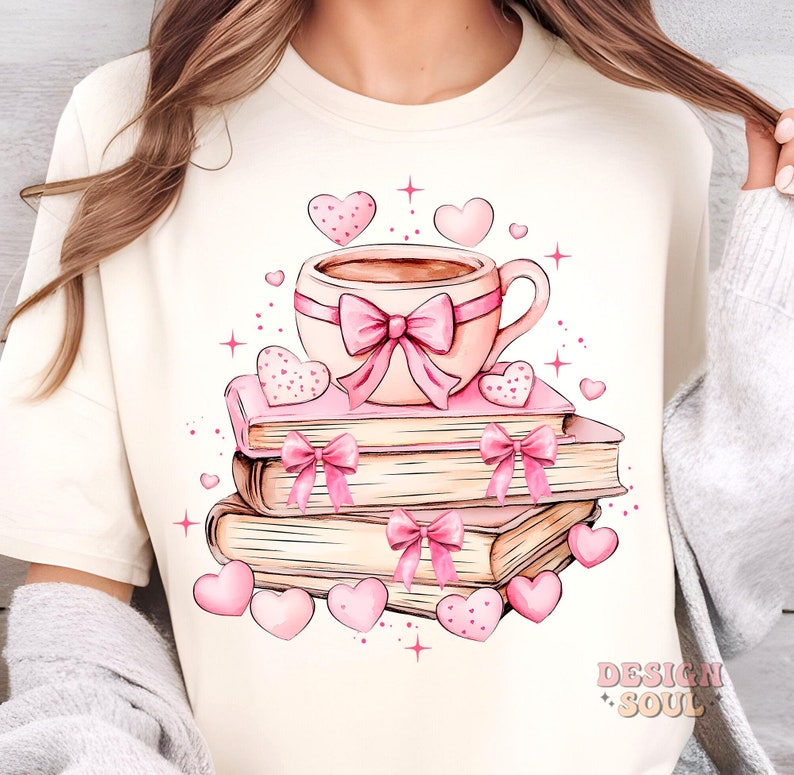 K&ouml;nnte beinhalten: Ein wei&szlig;es T-Shirt mit einer rosa Kaffeetasse, einer rosa Schleife und einem Stapel B&uuml;cher mit rosa Schleifen. Die B&uuml;cher sind von rosa Herzen und Sternen umgeben.