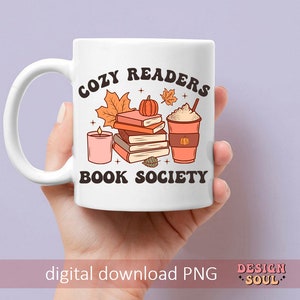 May include: White ceramic mug with a brown and orange design. The mug features a stack of books, a candle, a pumpkin, and a cup of coffee. The text on the mug reads "Cozy Readers Book Society".