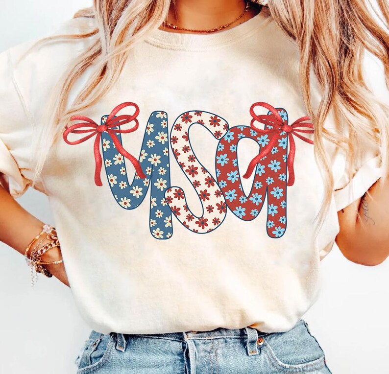 May include: A white t-shirt with a red, white, and blue floral design that spells out "USA" with red bows on each side of the letters.