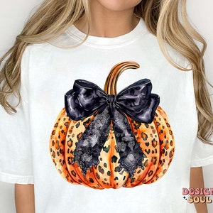 May include: White t-shirt with a graphic of a pumpkin with a black and white lace bow. The pumpkin is orange and black with a leopard print pattern.