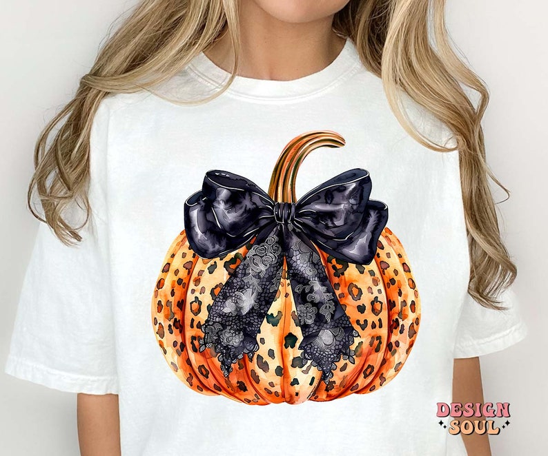 May include: White t-shirt with a graphic of a pumpkin with a black and white lace bow. The pumpkin is orange and black with a leopard print pattern.