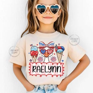 May include: A beige t-shirt featuring a patriotic design with ice cream cones, fireworks, and a duck wearing heart-shaped sunglasses. The name "RAELYNN" is displayed in a red and pink frame. The shirt is worn by a young girl.