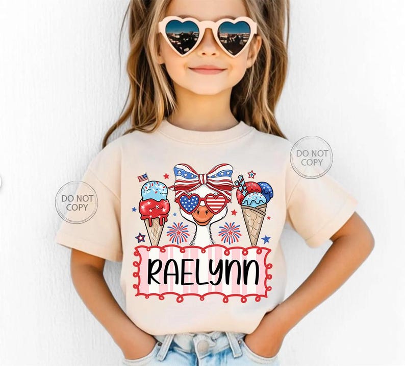 May include: A beige t-shirt featuring a patriotic design with ice cream cones, fireworks, and a duck wearing heart-shaped sunglasses. The name "RAELYNN" is displayed in a red and pink frame. The shirt is worn by a young girl.