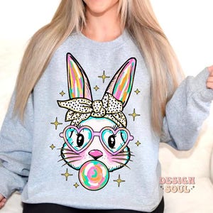 May include: A light gray sweatshirt with a colorful cartoon bunny wearing heart-shaped sunglasses and a bow. The bunny is holding a colorful lollipop. The text "DESIGN SOUL" is printed on the bottom right corner of the sweatshirt.